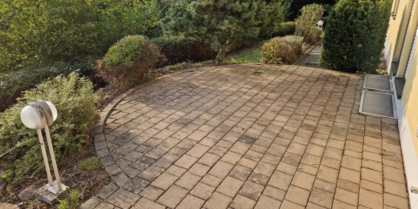 Gepflegte gepflasterte Terrasse mit runder Einfassung, umgeben von Sträuchern, Bäumen und Gartenbeleuchtung neben einem gelben Wohnhaus.