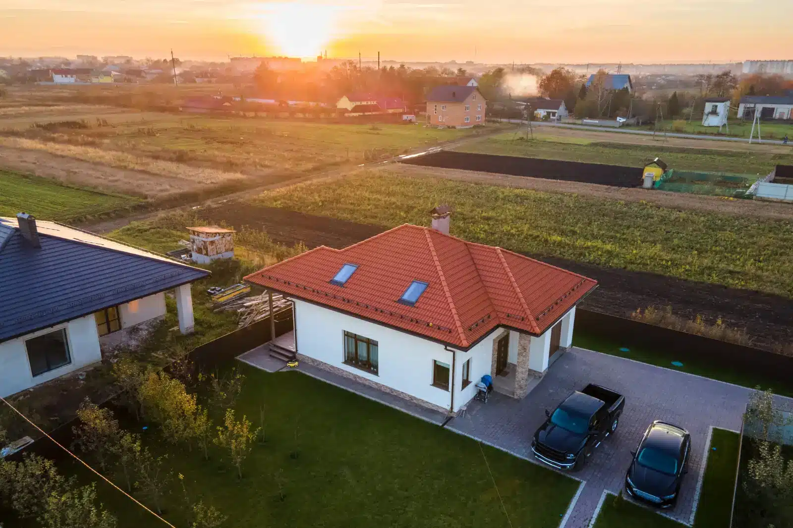 Modernes Einfamilienhaus mit rotem Ziegeldach und gepflegtem Garten in ländlicher Umgebung bei Sonnenuntergang, mit zwei geparkten Autos in der Einfahrt.
